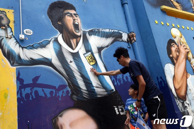 25일(현지시간) 부에노스아이레스 라보카 인근에서 아르헨티나의 축구 전설 디에고 마라도나를 묘사한 벽화 옆에서 한 남자와 아이가 헌화를 하고 있다. © AFP=뉴스1 © News1 이동원 기자